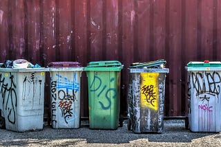 Color picture of five trashcans lined up against a corrgated iron wall