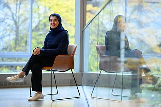 Sarah Alnegheimish sits in front of a wall of windows overlooking out-of-focus trees.