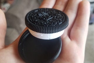 A black and white, 3D-printed Oreo box sits unscrewed in a white person’s hand. In the blurred background is a couch and floor.