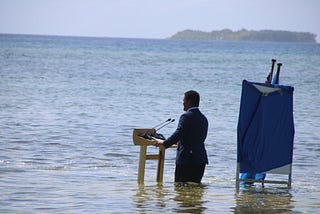 A man is standing knee-deep in water. In front of him is a desk with two microphones, behind him two flags.