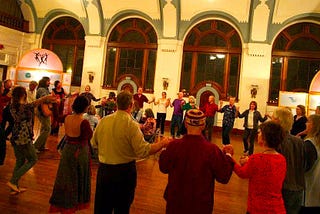 Dances of Universal Peace pre-pandemic. Perhaps 50 people in a circle in an old building with arched windows. Photo by Jennifer Stock by permission.
