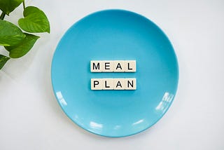 A blue plate displays letter tiles spelling out “MEAL PLAN” on a white background, accompanied by a leafy green plant to the side. The setup conveys themes of nutrition, organization, and healthy lifestyle planning. Reflecting the perspective of Ramneek Bhogal on how to support digestive resilience when traveling.