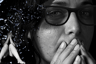 women with slight bruise near eye with tear over it hidden under dark sunglasses. In the corner under shattered glass a woman sits hiding her face.