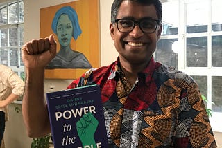 Danny, smiling, fist raised in ‘solidarity’ holding a copy of his book.