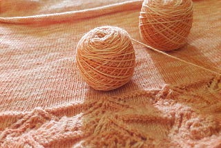 Two balls of pinkish yarn sit atop a sweater in progress, with knitting lace near the bottom.