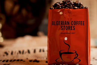 Image of Red Coffee Bag full of coffee beans — Bag printed “Algerian Coffee Stores Established 1887”, sitting on a coffee sack labelled SUMATRA