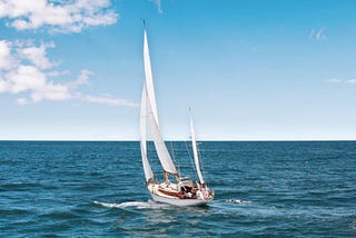 Blue ocean with sailboat with white clouds on left side of blue sky.