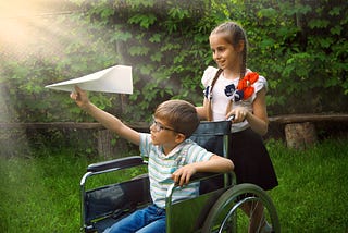 Girl with flowered blouse pushing a young boy in wheelchair, he is about to throw a paper airplane.