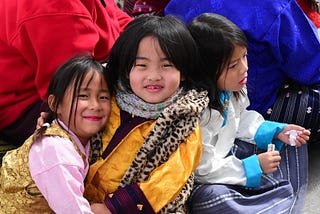Two small girls in traditional Bhutanese attire at a festival, smiling at the camera.