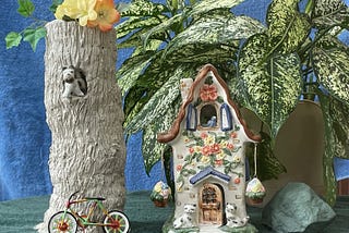 A clay tree trunk has a little ceramic dog standing inside a hole in it, a bicycle leaning against  it, and a colorful ceramic cottage next to it with a green plant and blue sky in the background.