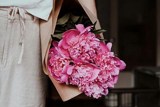 Woman carrying pink peonies wrapped in brown paper.