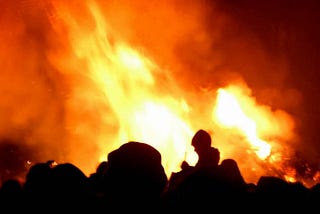 A crowd of people in silhouette, standing in front of a raging bonfire.