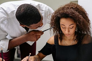 Man in a white shirt bends at the waist, leaning close to a woman and points a pencil at her face while she’s sitting at a desk.