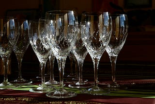 Crystal wineglasses are arranged on dark wood over a deep green table scarf in shadow with light shining on them at an angle.
