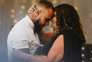 A woman in a black gown embraces a tattooed man in a white shirt
