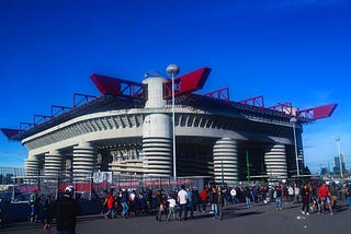 People outside a wire fence surrounding a stadium with large black and white striped pillars and red girders along the roof.