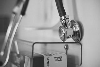 A black and white photo of a stethoscope hanging on a wall in a nondescript healthcare setting.