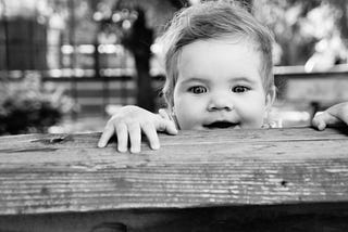 A black-and-white image of a young child peaking over the edge of a wooden beam, their hands reaching out inquisitively with a large smile on their face.