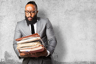 A photo of a man looking apprehensively at a large pile of folders and papers that he’s holding.