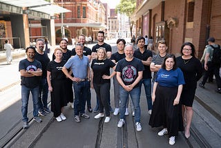 A group of people standing on tram tracks in front of red brick buildings, smiling. CyRise Accelerator startup founders with CyRise team. Back: Ron Moritz, Simon O’Keefe, Sam Stewart, Ivan Vanderbyl, Tony Mao, Shan Kulkarni, Tim Thacker and Fiona Long. Front: Tinesh Chhaya, Kirstin McIntosh, Lorenzo Modesto, Lainie Vinikoor, Scott Handsaker and Jenny Binetter