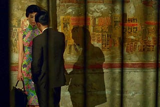 A man and woman stand together in front of a textured wall, engaged in conversation.