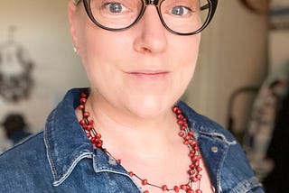 Woman with chemo hair growing out, wearing glasses, a red necklace and a denim jacket