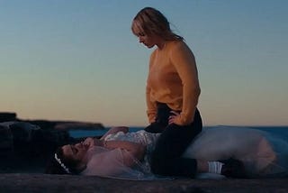 On a clifftop, a woman sits on top of another woman. The one underneath wears a wedding dress.