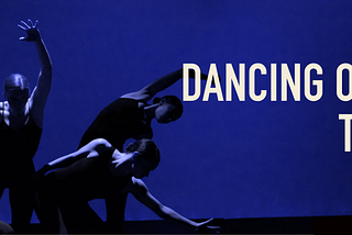 Dancers in low lighting, wearing black unitards, reach in different directions and intertwine their bodies.