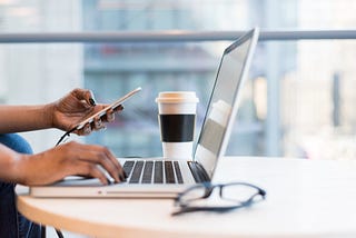 One hand on a laptop keyboard and other hand holding a phone. In addition to the laptop being on the table, there is also drink cup and a pair of glasses.