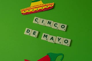 Cinco de Mayo celebration decorations on a bright green background featuring letter tiles spelling ‘CINCO DE MAYO’, a colorful sombrero cutout, and a red chili pepper cutout.