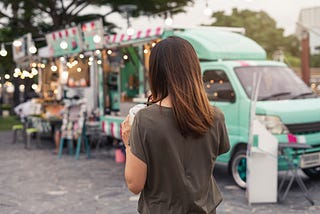 Image of a person with long brown hair and a gray shirt sipping out of a paper cup in front of a mint green food truck display