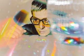 Photograph of a white, non-binary person wearing glasses with short hair. They are resting their chin on a table and look frustrated. The photo is abstract with blurry swipes, dots and other shapes overlaid on the image.