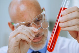 Photo of an older man in a lab coat examining a test tube filled with red liquid using a magnifying glass. That is to say, there is no actual science taking place in the photo, just the veneer of “science-ness.”