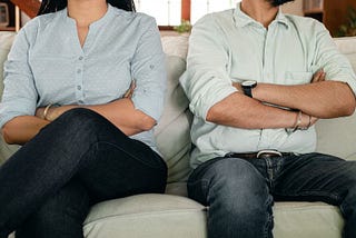 A man and woman are seated and can only be seen from the waist down. They are both wearing a light gray shirt and blue jeans.