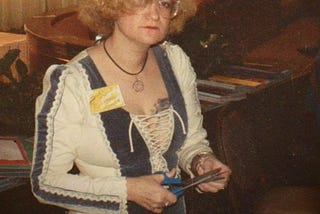 Photo is of a young blonde woman with curly neck-length hair, a feathered headband, and glasses, wearing a 1970’s Renaissance-style maxi dress in blue velvet and cream colored linen. She stand behind a table and holds a pair of scissors. There are papers and pamphlets on the table before her and behind her.