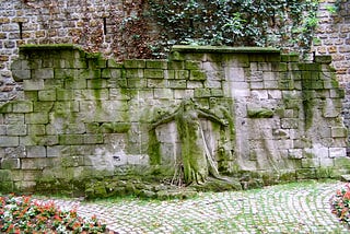 Carved in relief on an old stone wall is a plaintive female figure, arms outreached, eyes skyward, who can be clearly seen. Less visible are the much shallower reliefs, the carvings of people, mostly men with mustaches, who seem to hover around her.