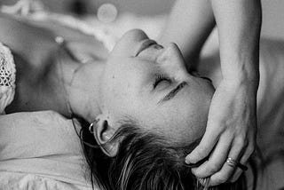 A woman lays back on her bed, clutching her forehead, her eyes are closed. The image is grayscale.