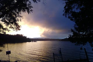 Bright and dark clouds over calm waters with overhanging cedar branches in the foreground.