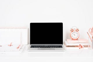 A Macbook, notebooks and a rose gold clock on a white desk