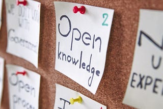 Yellow post-it notes attached to a cork board with colorful push-pins. The fully visible notes say (from ascending to descending), “Education ? ? ?,” “Open Knowledge,” and “Team” crossed out with “Community” written below.