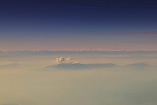 A deep blue sky above wispy white clouds, as if seen from an aeroplane. Mountain tops can just be seen through the clouds in the distance.