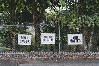 Chain link fence with three white signs with black letters saying Don’t Give Up, You Are Not Alone, You Matter.