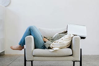 A woman lying sideways in an armchair with an open notebook covering her face.