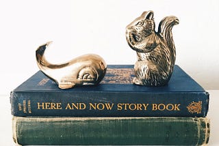 A pile of two books lay flat on a white surface. The bottom book is green with worn edges. There is no title on the spine. The second book lies on top of the green one. It’s blue with worn edges and gold lettering. It reads ‘Here and now story book’. There is also an insignia and other writing, which is too small to decipher. On top of the books are a brass whale and squirrel.