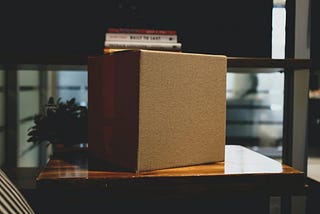 A plain corrugated box sits on top of a desk. A stack of books and plant can be spotted in the darkened background.