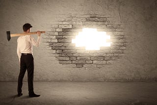 a man with a sledge hammer stands in front of a brick wall. He has knocked a small hole in the wall and sunlight is shining through.