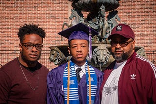 2 young black men and their father. One of the young men is in graduation attire.