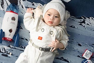 An infant wears a white spacesuit-looking sleeper and lays against a surface-of-the-moon themed blanket. A toy rocket lies on its side next to the baby.