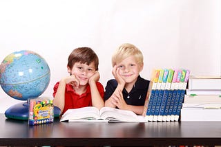 Two children sitting and smiling with textbooks, a globe, and some markers