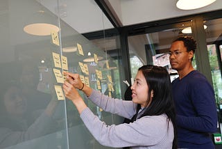 A woman in a light blue sweater places a sticky note on an idea board. A man with glasses stands beside her.
